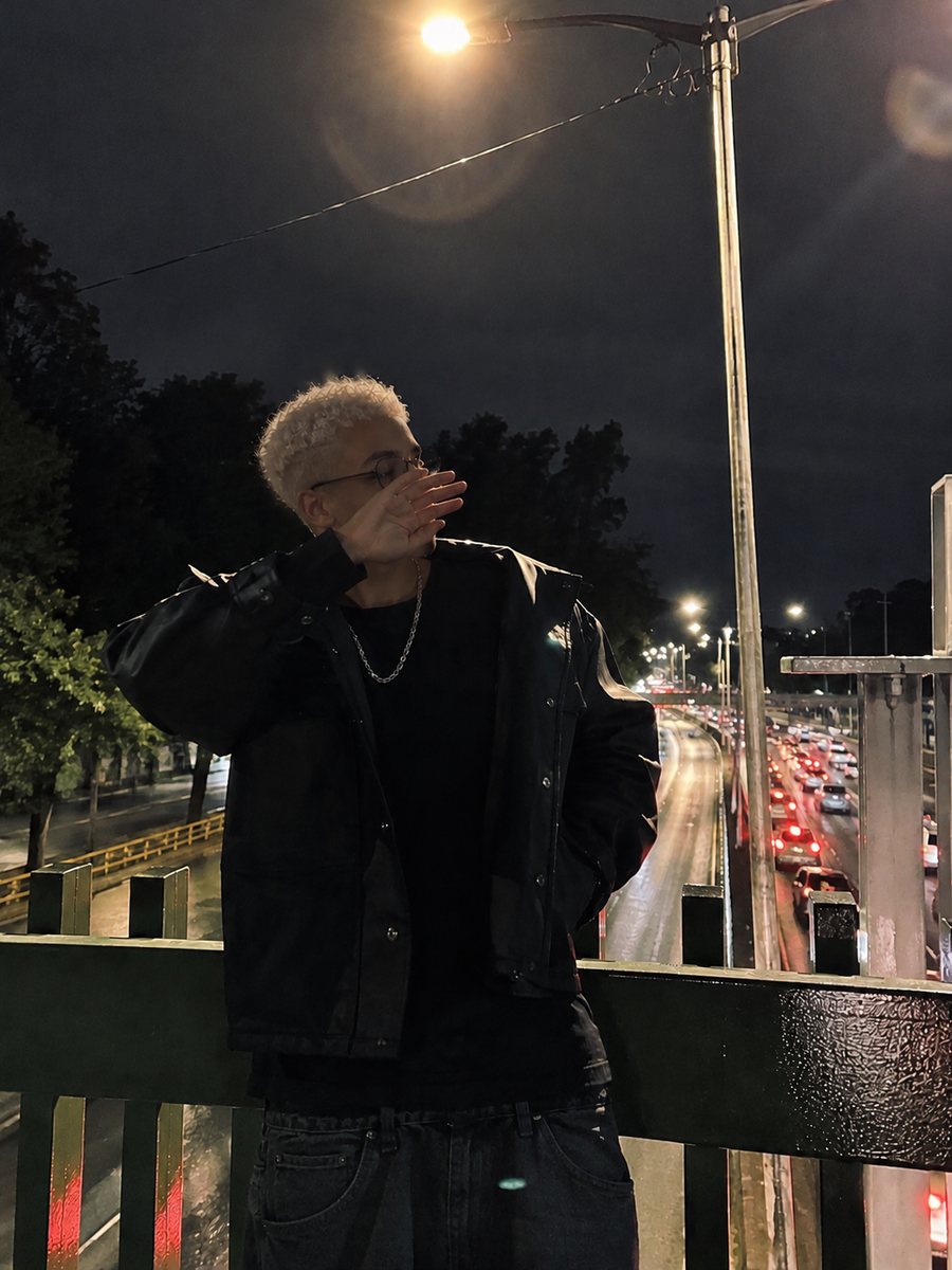 Igor at night on a footbridge over a busy avenue, wearing a black jacket, with a hand in front of his face.